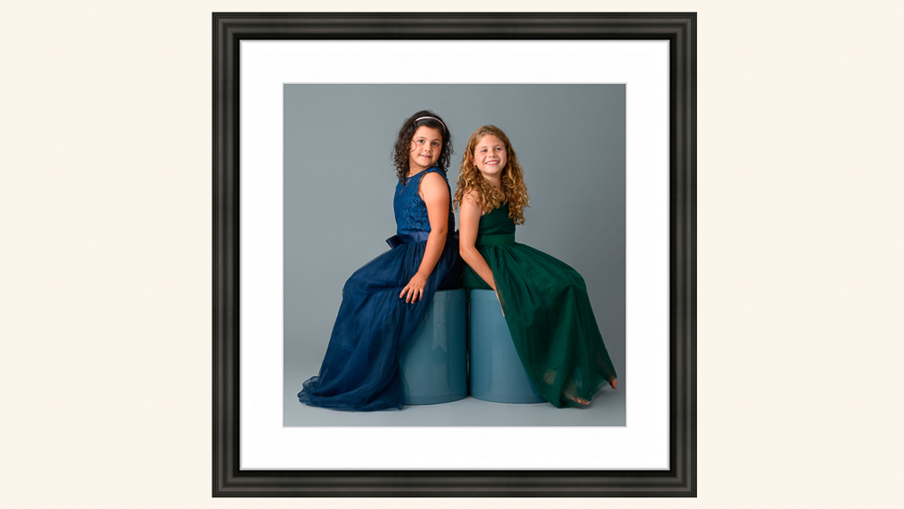 Two young girls in elegant ball gowns, one wearing blue and one in green, posed back-to-back against a gray studio background | Lisa Maco Photography