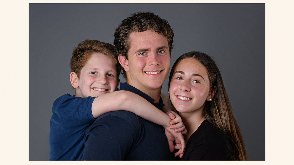 Three smiling siblings posing together against a gray background in a studio portrait | Lisa Maco Photography