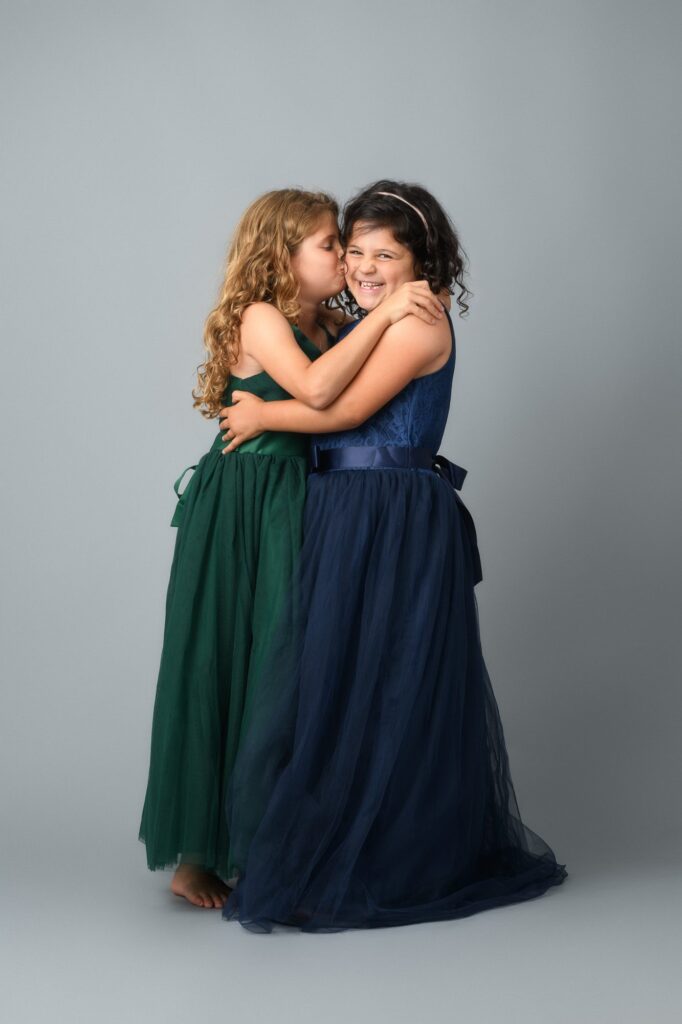 Two young girls embrace and smile during a DC family studio portrait session, wearing flowing green and navy dresses against a neutral gray background.