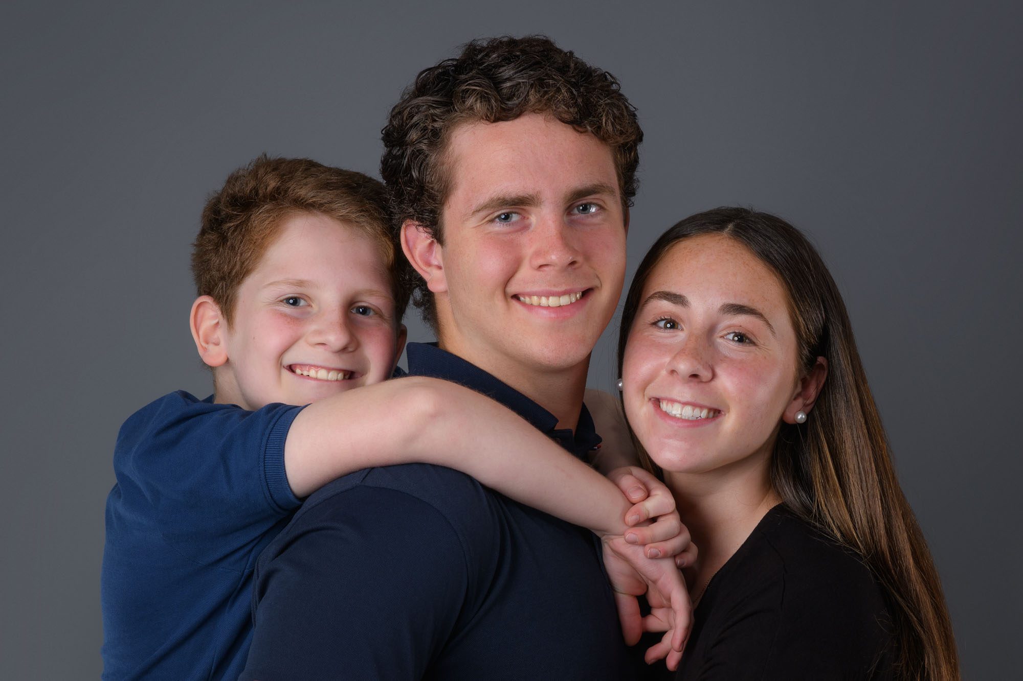 Three smiling siblings pose together during a Washington DC family portrait session, capturing the warmth and connection of sibling bonds in a studio setting.