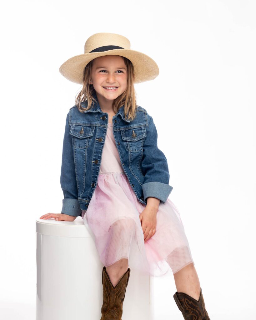Young girl smiling during a studio family photography session, wearing a denim jacket, pink tulle skirt, and cowboy boots with a straw hat
