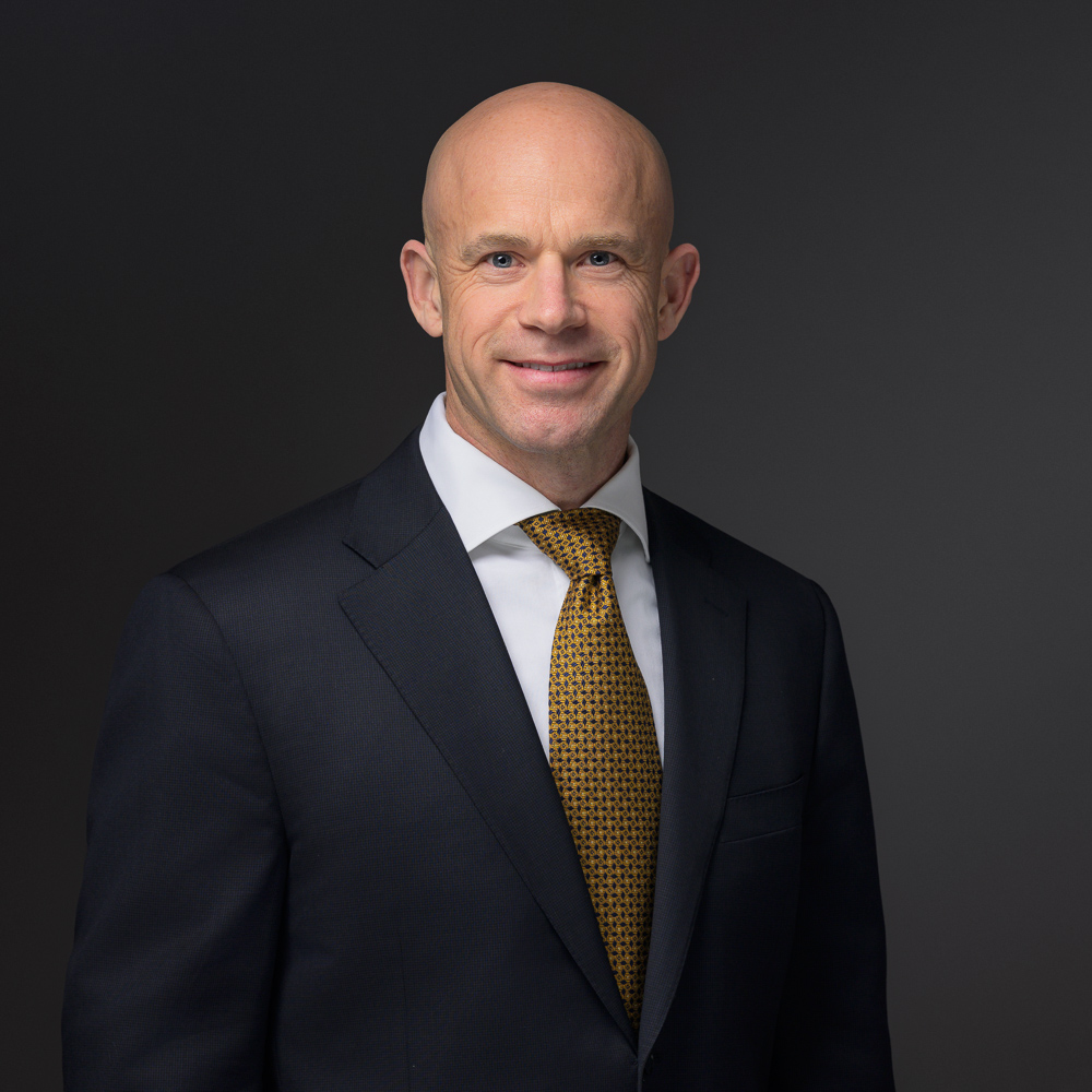 Professional headshot of a bald man in a dark suit, white dress shirt, and gold patterned tie against a dark gray background