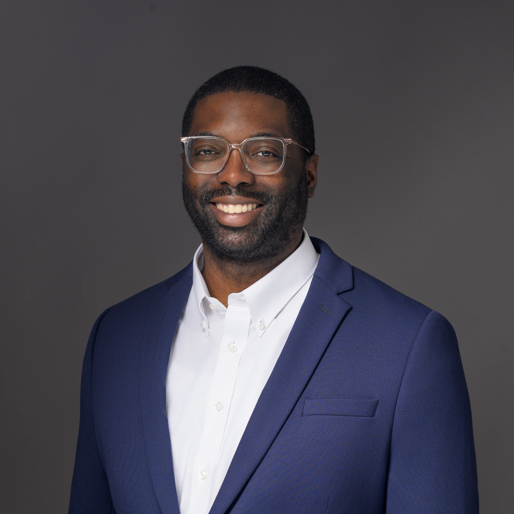 Professional headshot of a man wearing clear-framed glasses, navy blazer, and white button-up shirt against a gray background