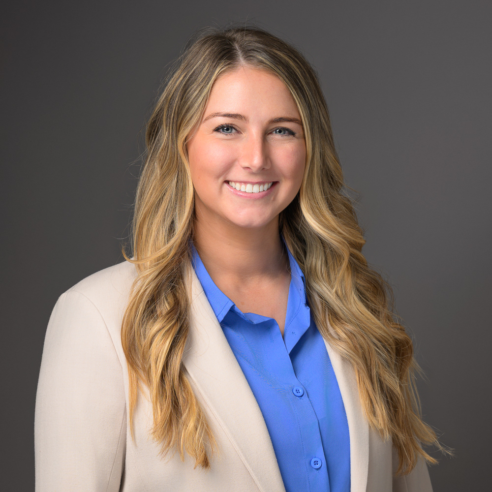 Professional headshot of a smiling woman with long brown hair wearing a blue blouse and cream blazer against a gray background