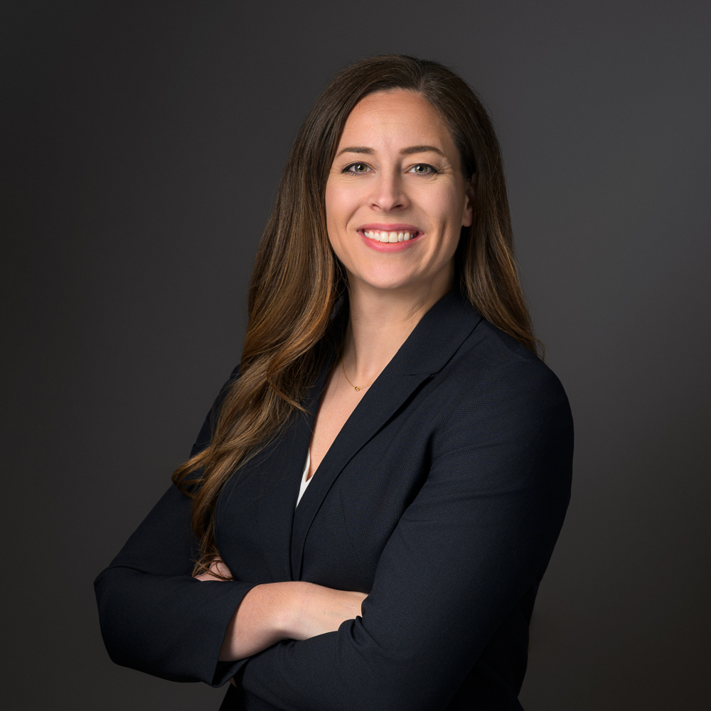 Professional woman with long brown hair smiling at camera wearing dark blazer against gray background