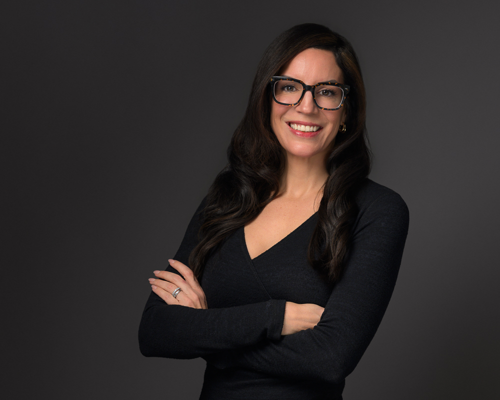 Professional headshot of a smiling woman with long dark hair and glasses wearing a black v-neck top