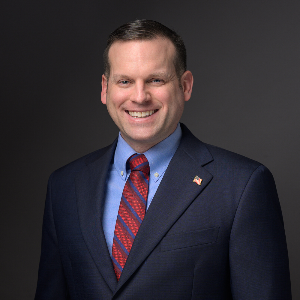 Professional headshot of smiling man in navy blazer and red striped tie against gray background