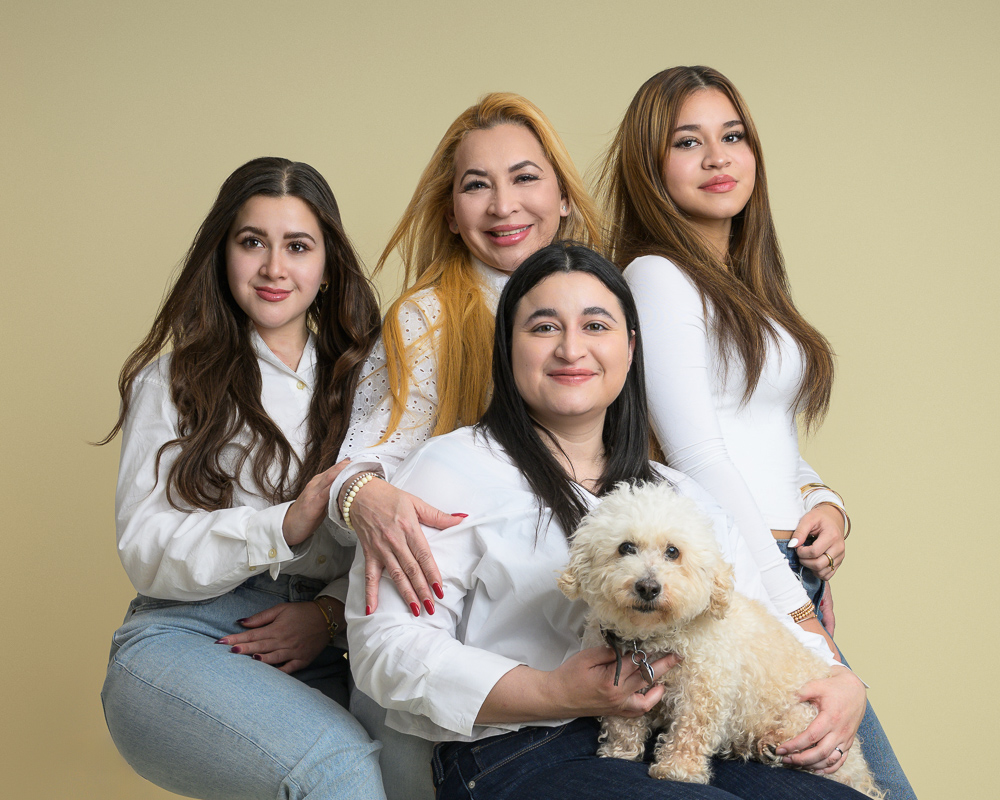 Four women and a cream-colored dog posing together against a beige background for a family portrait