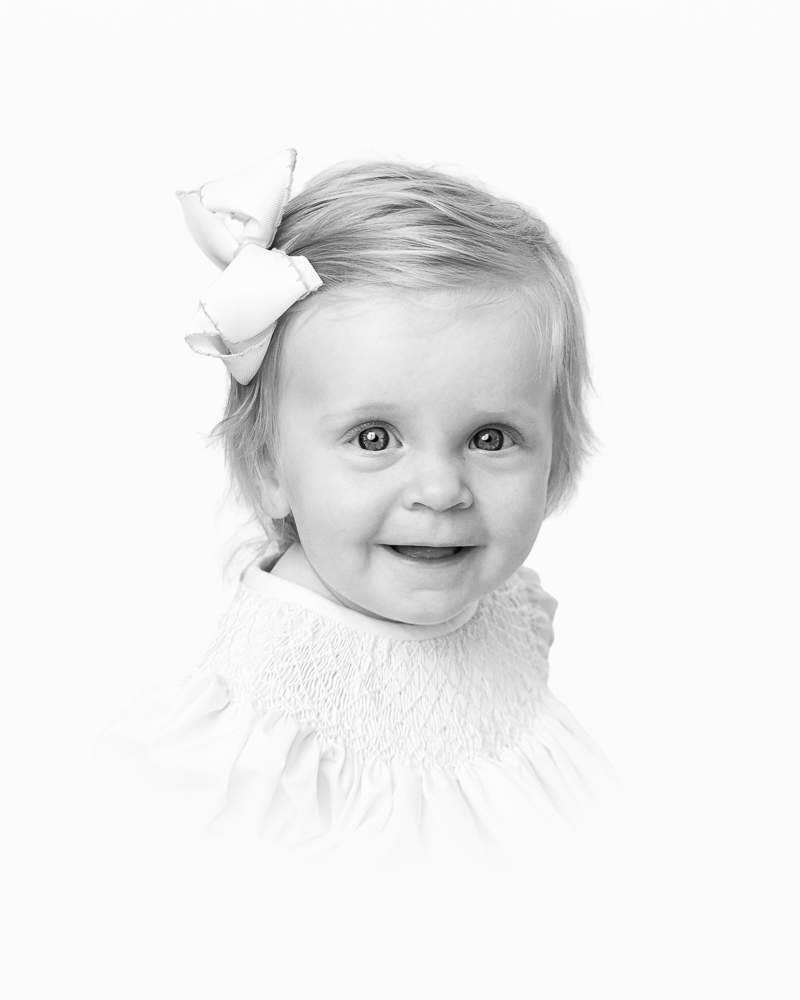 Smiling toddler with a white bow in her hair, captured in a classic black and white studio portrait by a DC family photographer