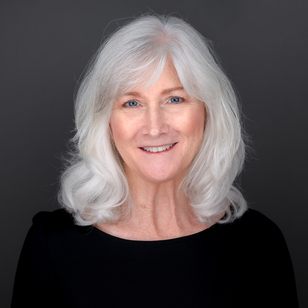 Professional headshot of a smiling woman with shoulder-length silver hair wearing a black top against a gray background