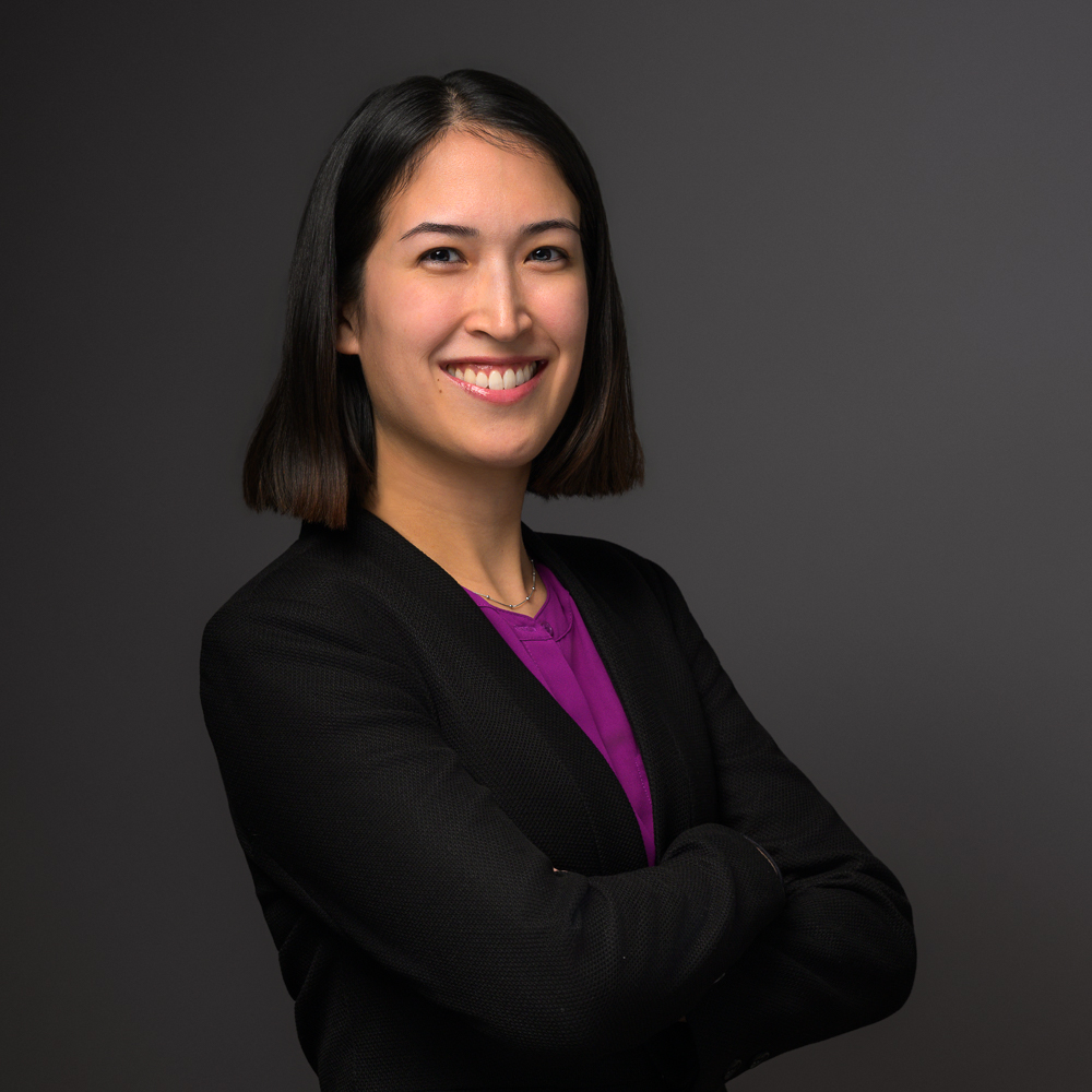 Professional headshot of smiling woman with shoulder-length black hair wearing black blazer and purple top against gray background