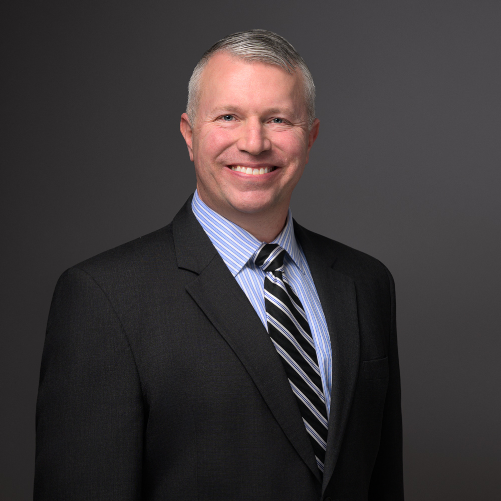 Professional headshot of a smiling man in a black blazer, light blue striped shirt, and dark striped tie against a gray background