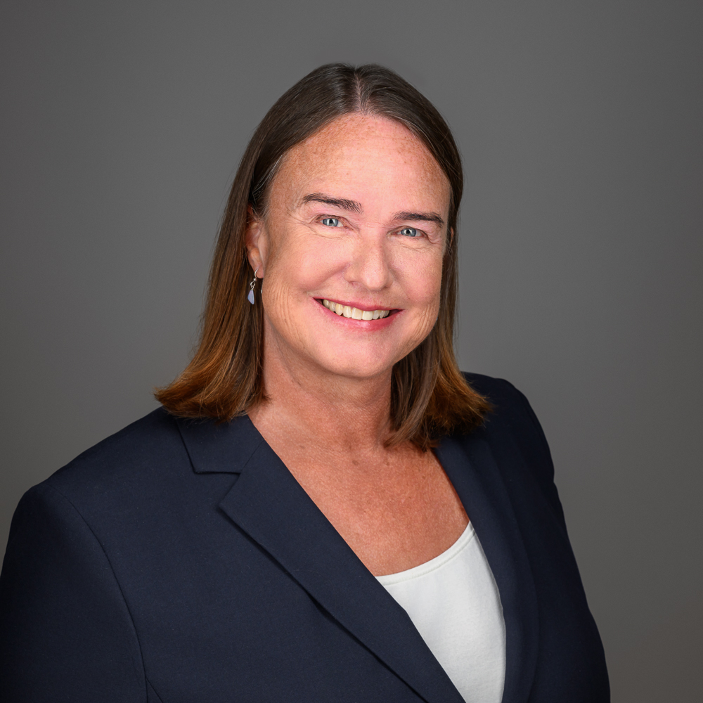 Professional headshot of a smiling woman with long brown hair wearing a dark blazer over a white top