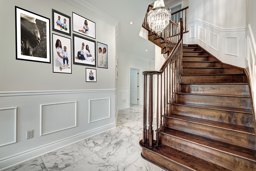 Elegant hallway with framed family portraits added over time on white paneled wall and wooden staircase with crystal chandelier